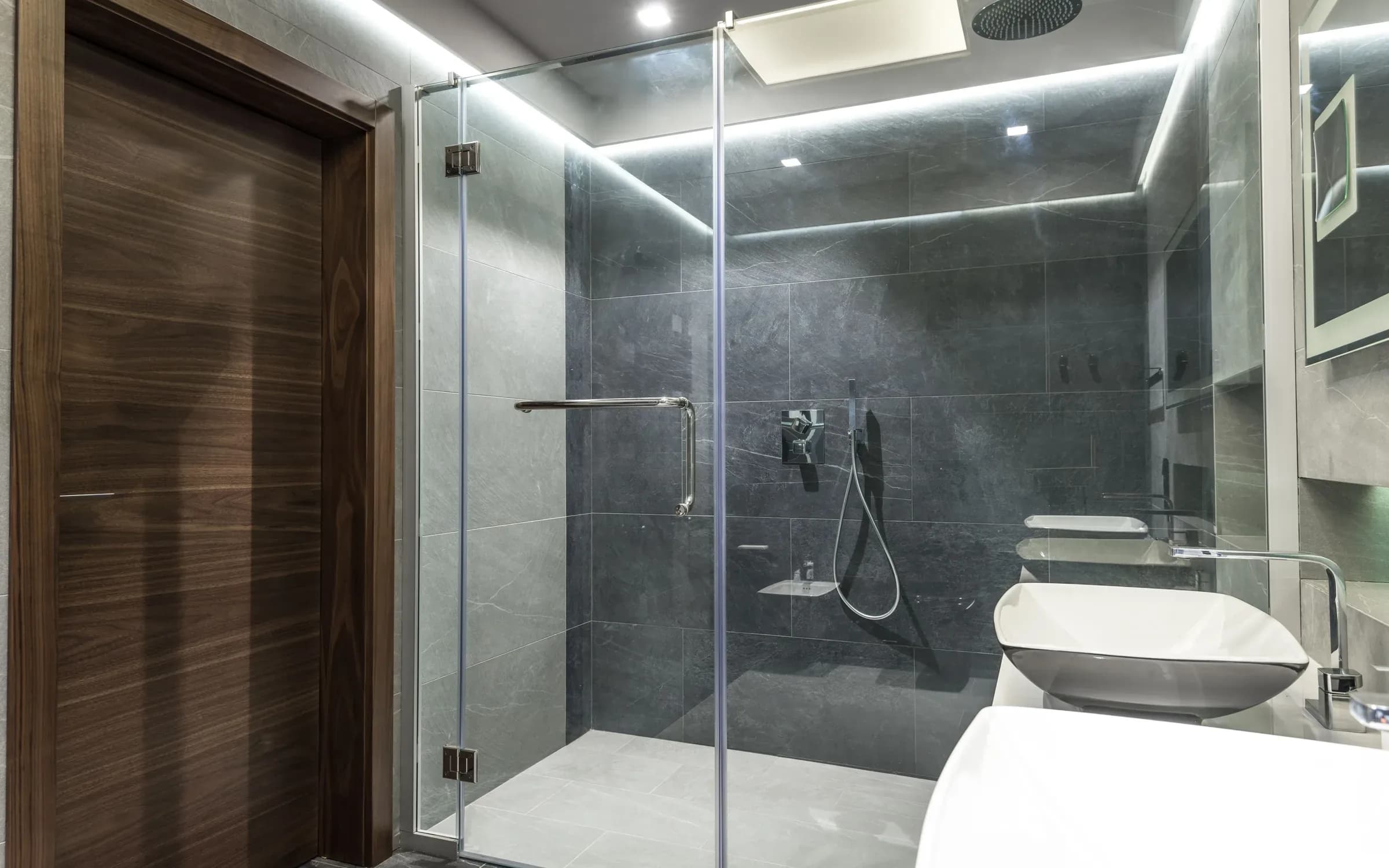 Koyu gri seramik, cam duş ve duvar tipi lavabo düzeni bulunan modern banyo